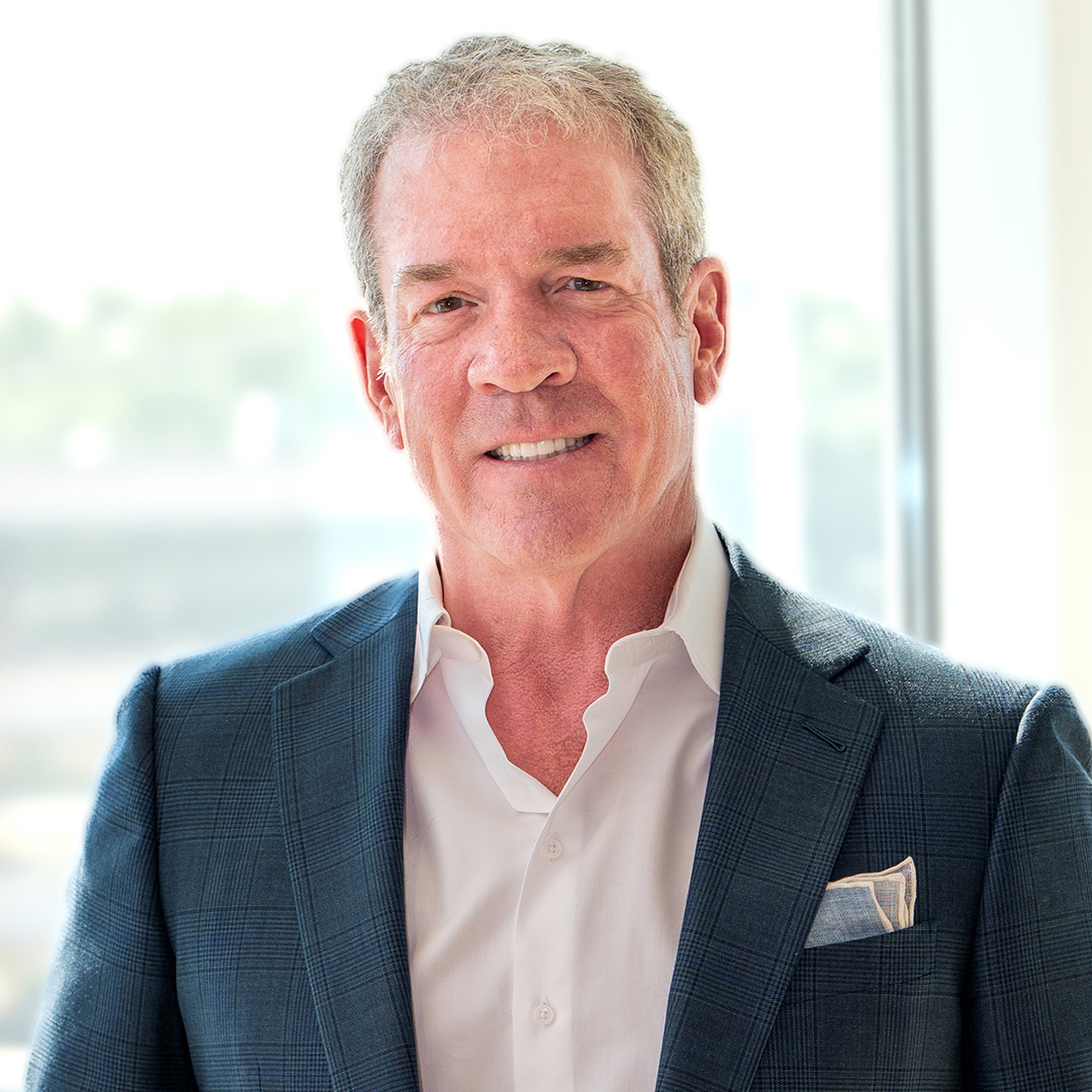 A man with gray hair wearing a blue suit jacket, white shirt, and pocket square stands in front of a bright window.
