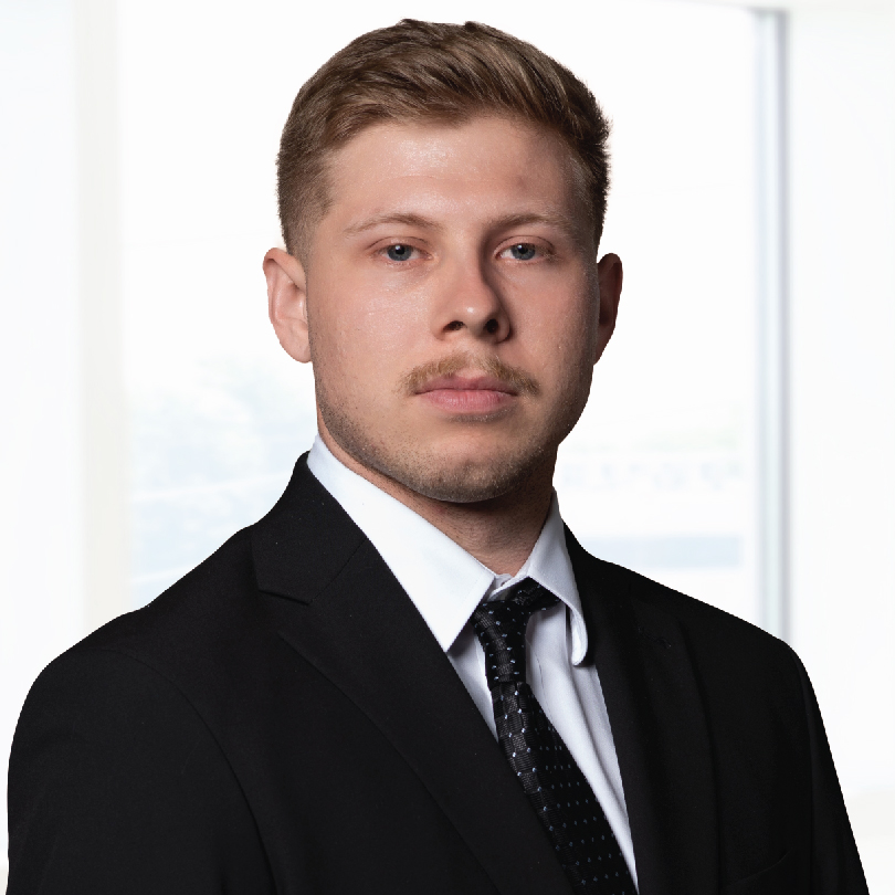 A man with short blond hair, wearing a black suit, white shirt, and black tie, stands in front of a bright window.