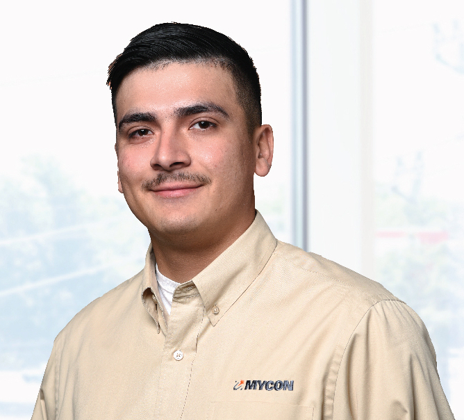 A person with short dark hair and a mustache wears a beige shirt with "MYCON" embroidered, standing indoors near a large window.