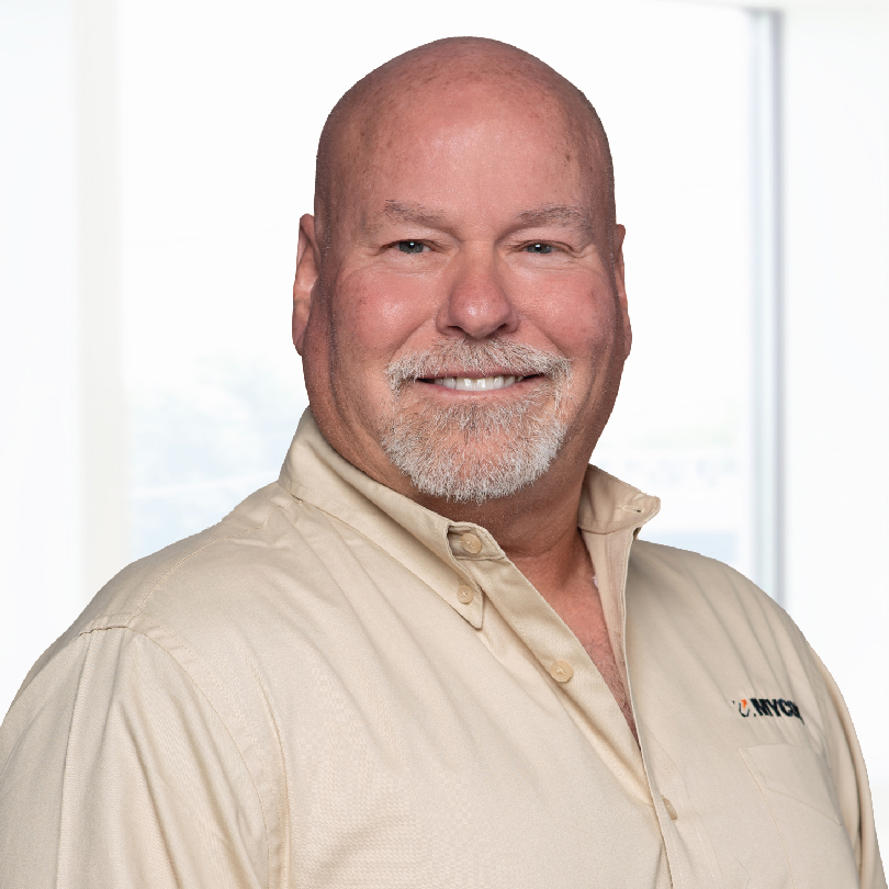 A smiling man with a shaved head and white beard is wearing a beige collared shirt in front of a light background.