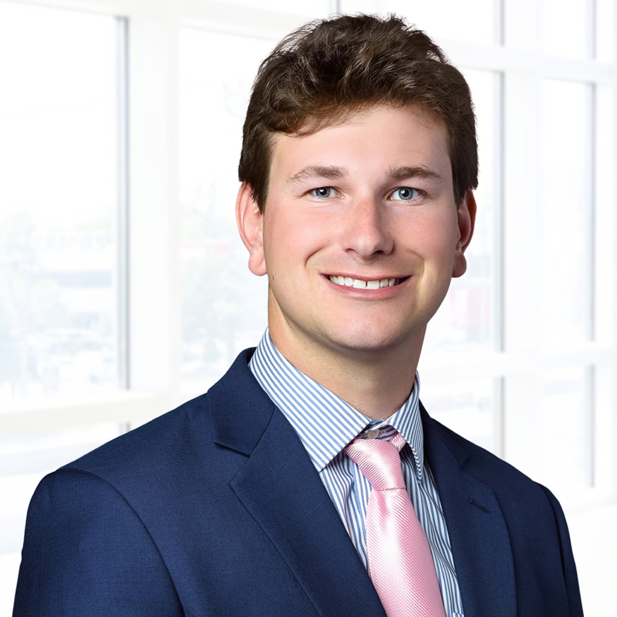 A man in a blue suit and pink tie stands in front of large windows, smiling at the camera.