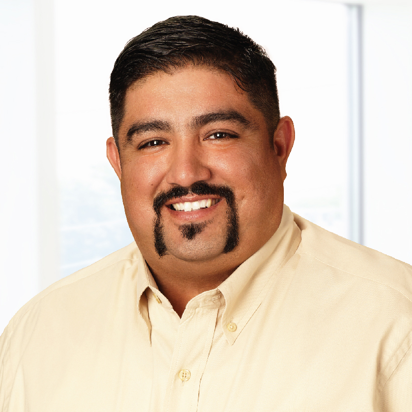 Man with short dark hair, mustache, and goatee, wearing a beige collared shirt, smiling in front of a bright, blurred indoor background.