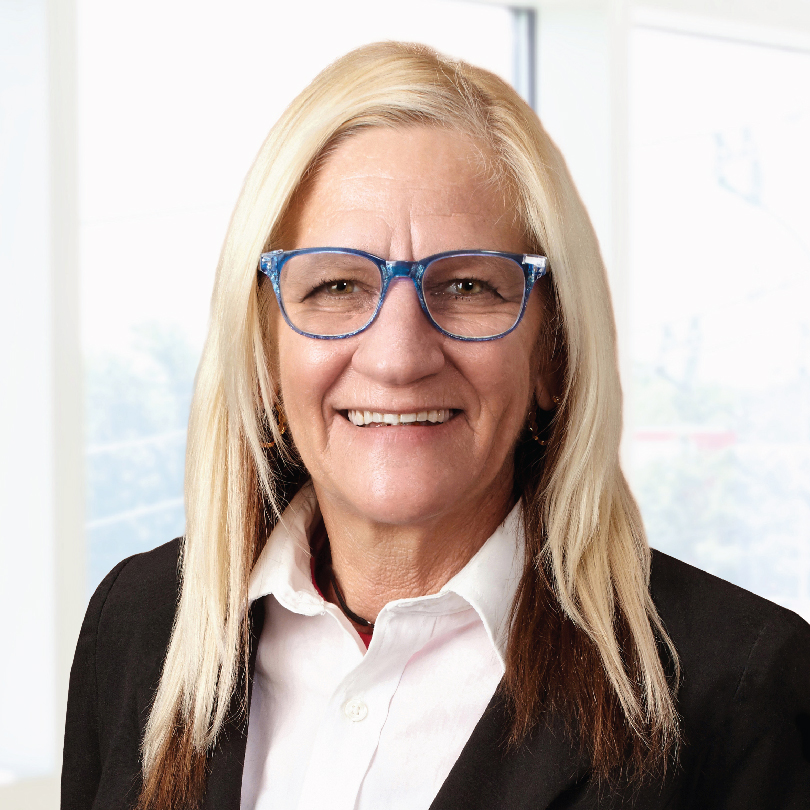 A person with long blonde hair and blue glasses wearing a white shirt and black blazer stands in front of a brightly lit window.