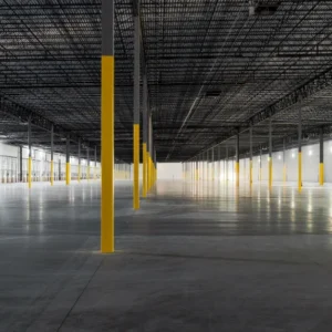 An empty, spacious warehouse interior at the Greenmount Logistics Center features concrete floors and rows of yellow-painted support columns under strategic overhead lighting, setting the stage for future construction innovations.