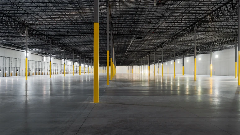 An empty, spacious warehouse interior at the Greenmount Logistics Center features concrete floors and rows of yellow-painted support columns under strategic overhead lighting, setting the stage for future construction innovations.