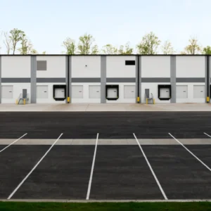 An empty parking lot stretches before the expansive Greenmount Logistics Center, with its multiple loading dock doors awaiting use. The scene is framed by trees in the background, highlighting the area's ongoing construction development.