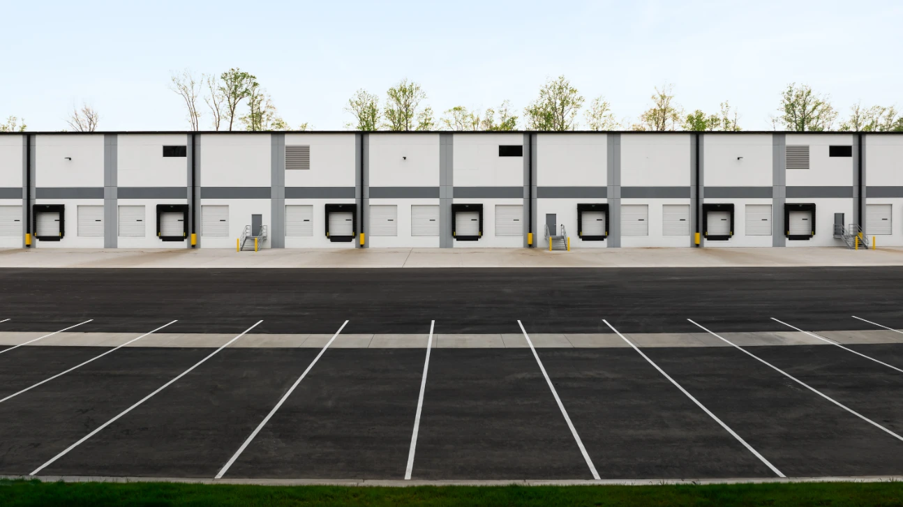 An empty parking lot stretches before the expansive Greenmount Logistics Center, with its multiple loading dock doors awaiting use. The scene is framed by trees in the background, highlighting the area's ongoing construction development.