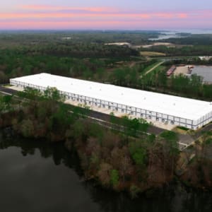 Aerial view of the expansive Greenmount Logistics Center under construction, its large white warehouse harmoniously nestled among trees beside a serene body of water, all enveloped by a cloudy sunset sky.