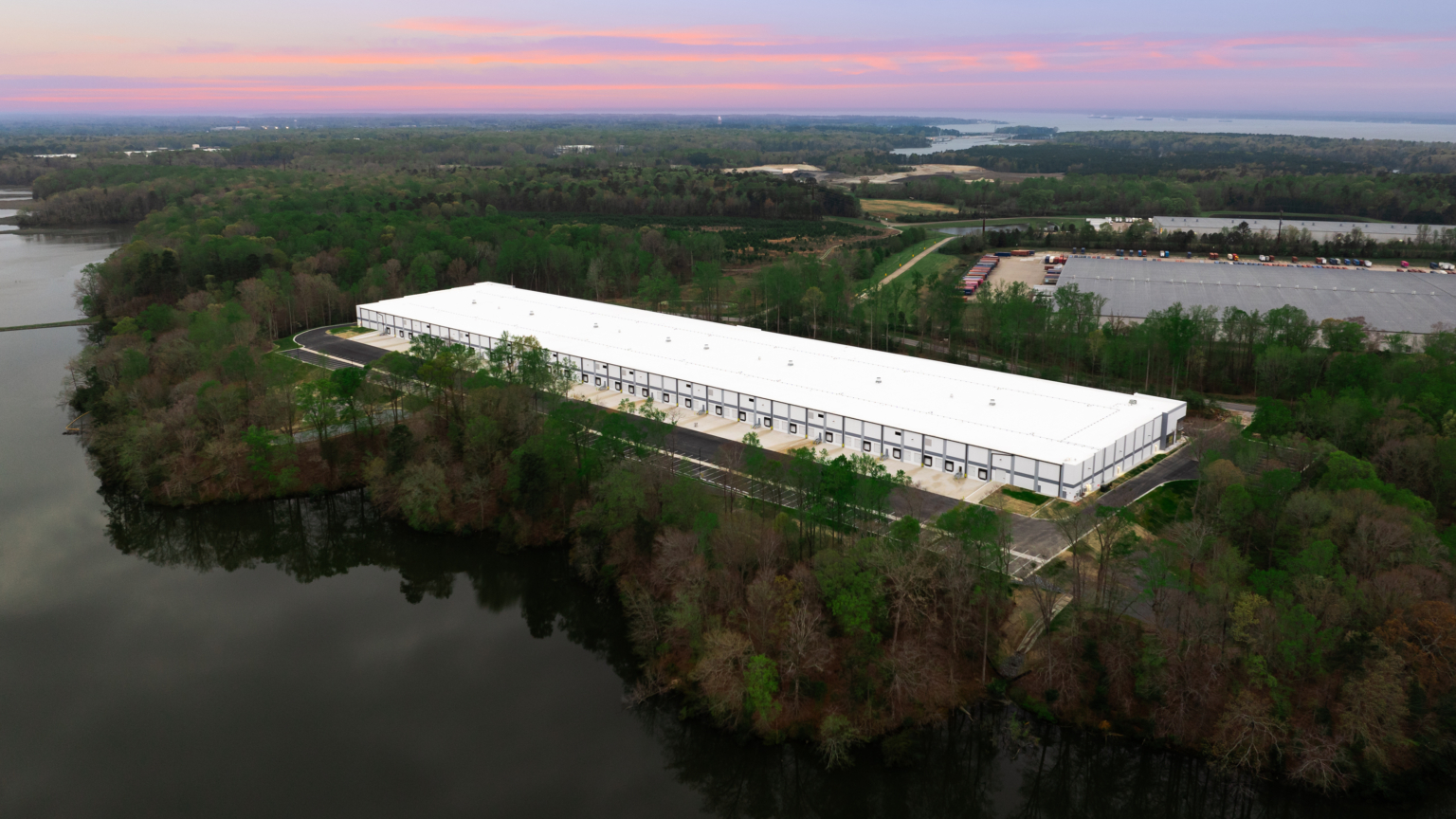 Aerial view of the expansive Greenmount Logistics Center under construction, its large white warehouse harmoniously nestled among trees beside a serene body of water, all enveloped by a cloudy sunset sky.