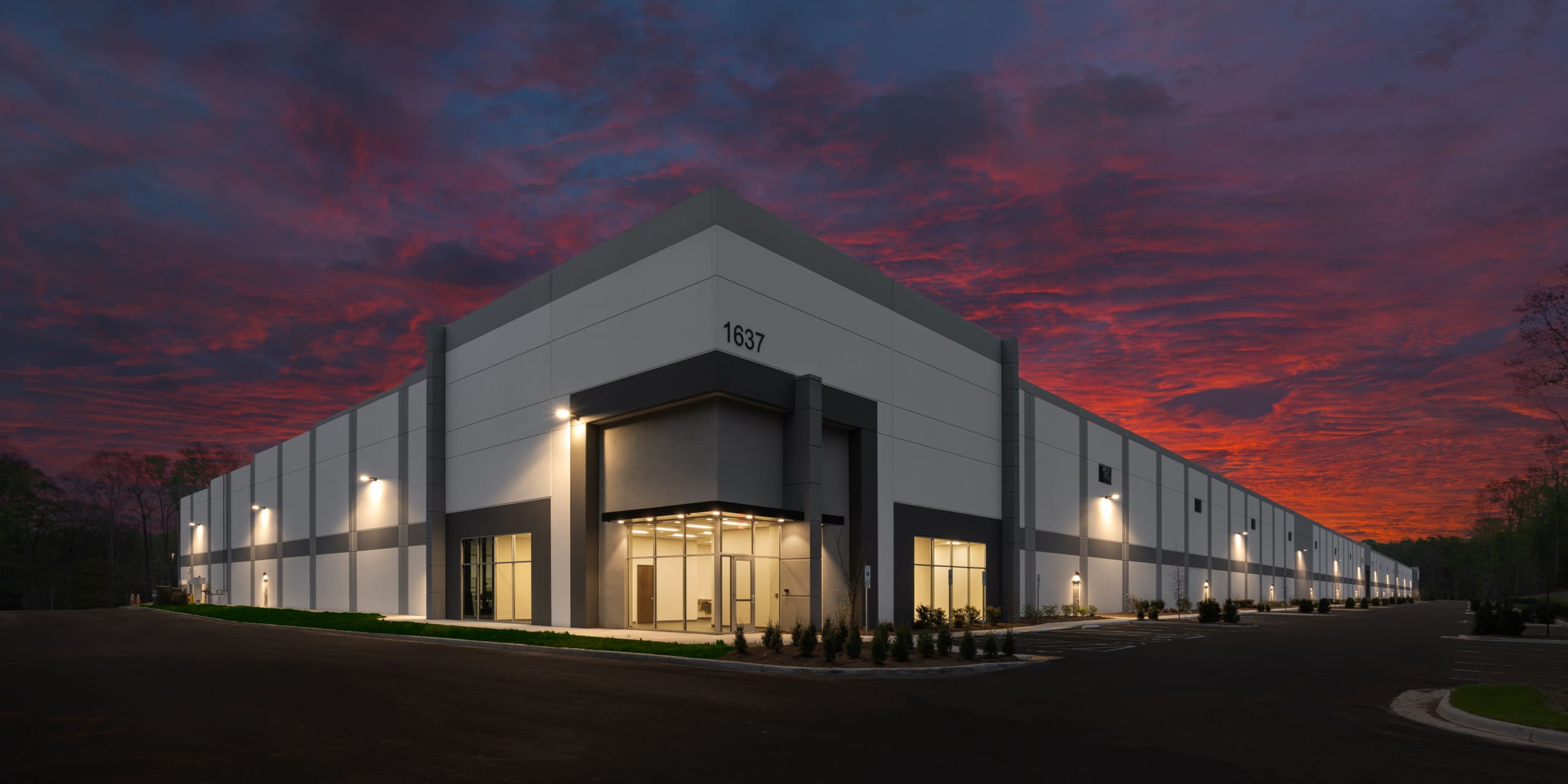 Modern industrial warehouse building photographed at dusk with illuminated exterior and vibrant red sky.
