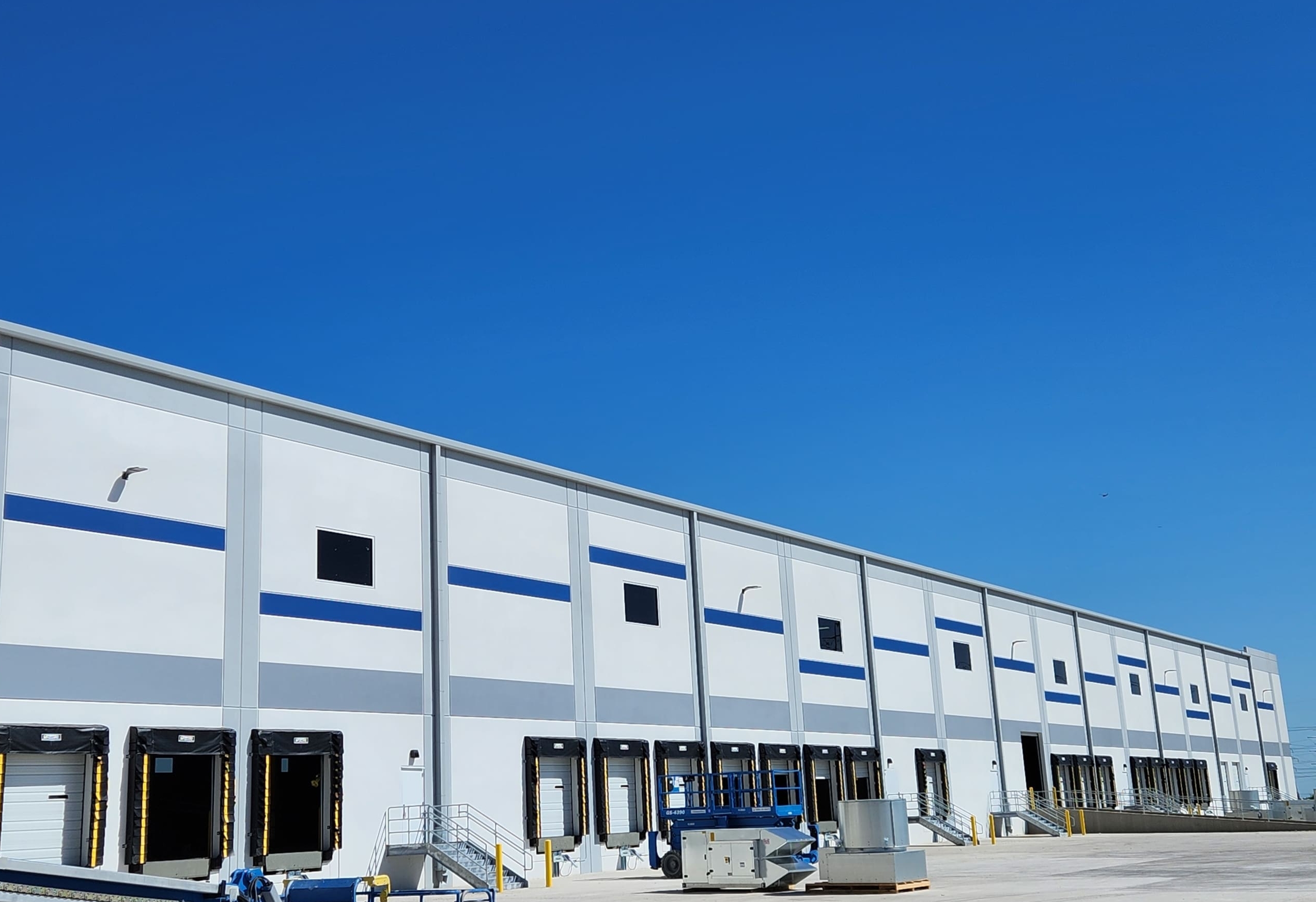 A modern industrial warehouse with a white facade and blue accents. Multiple loading docks along the side of the building are visible under a clear blue sky.