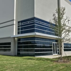 A large modern commercial building with a glass corner entrance, white and gray exterior, and a small grassy area with a few young trees in front.