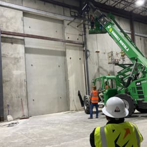 Two construction workers standing next to a crane in a warehouse.
