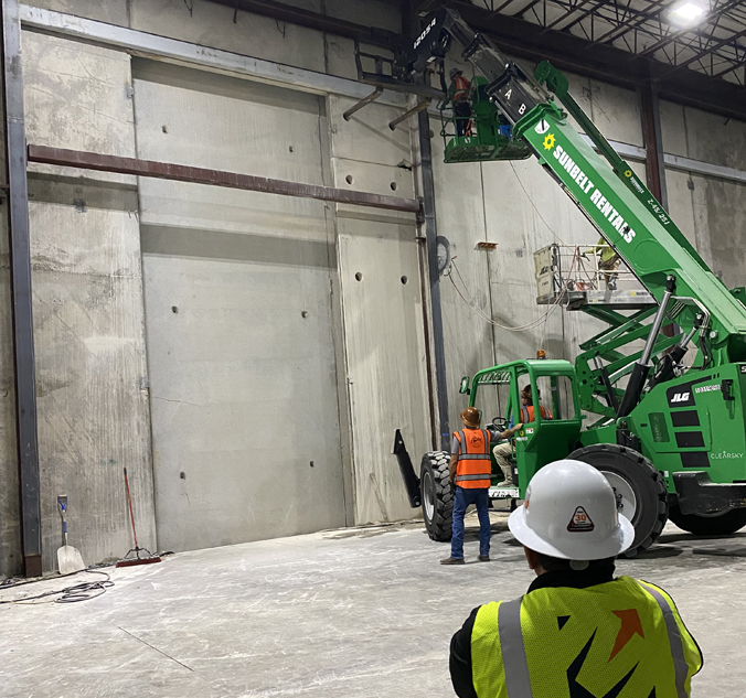 Two construction workers standing next to a crane in a warehouse.