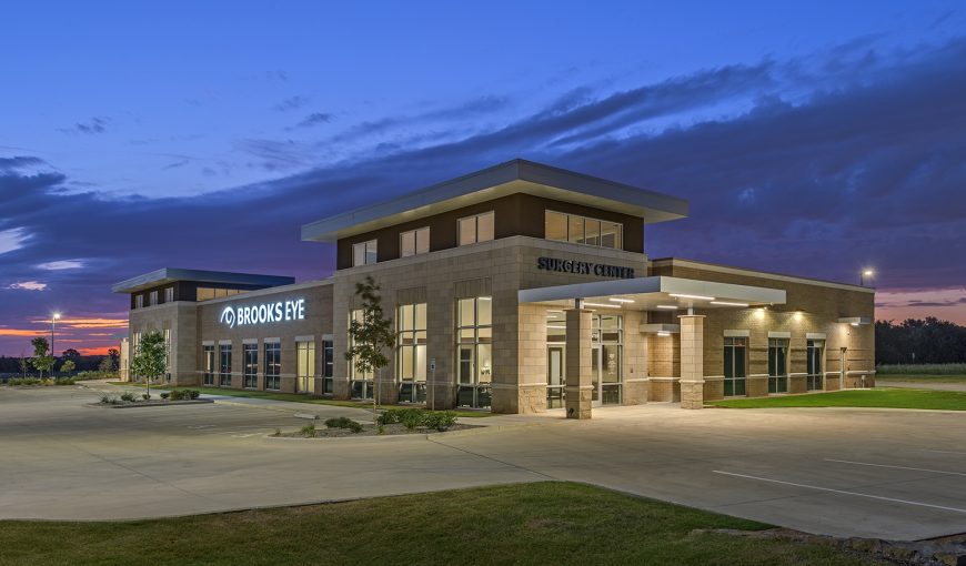 A modern, single-story medical building labeled "Brooks Eye Surgery Center" at dusk, with exterior lights on and an empty parking lot in front, representing the Brooks Eye Associates Ambulatory Surgery Center.