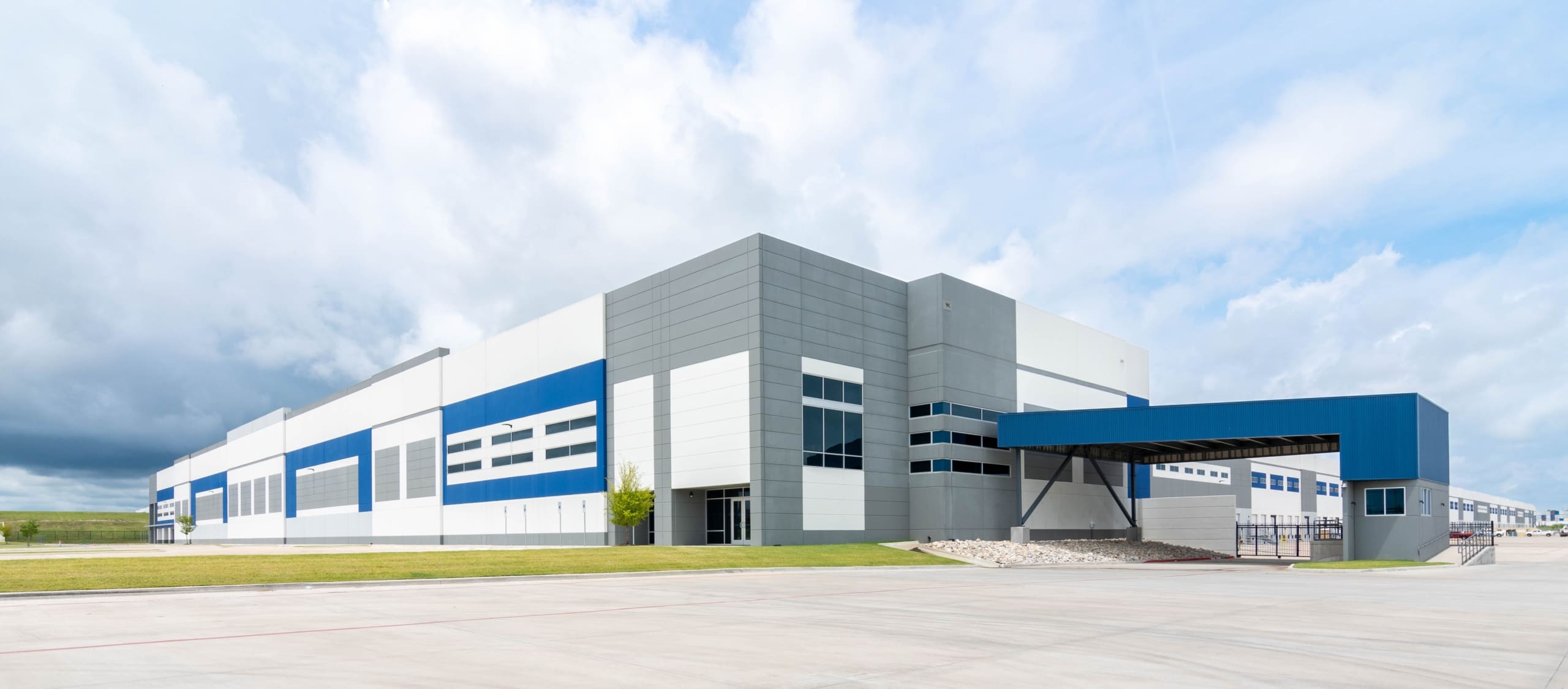 Large modern industrial warehouse building with white and gray exterior, blue accents, and loading docks, set under a partly cloudy sky.
