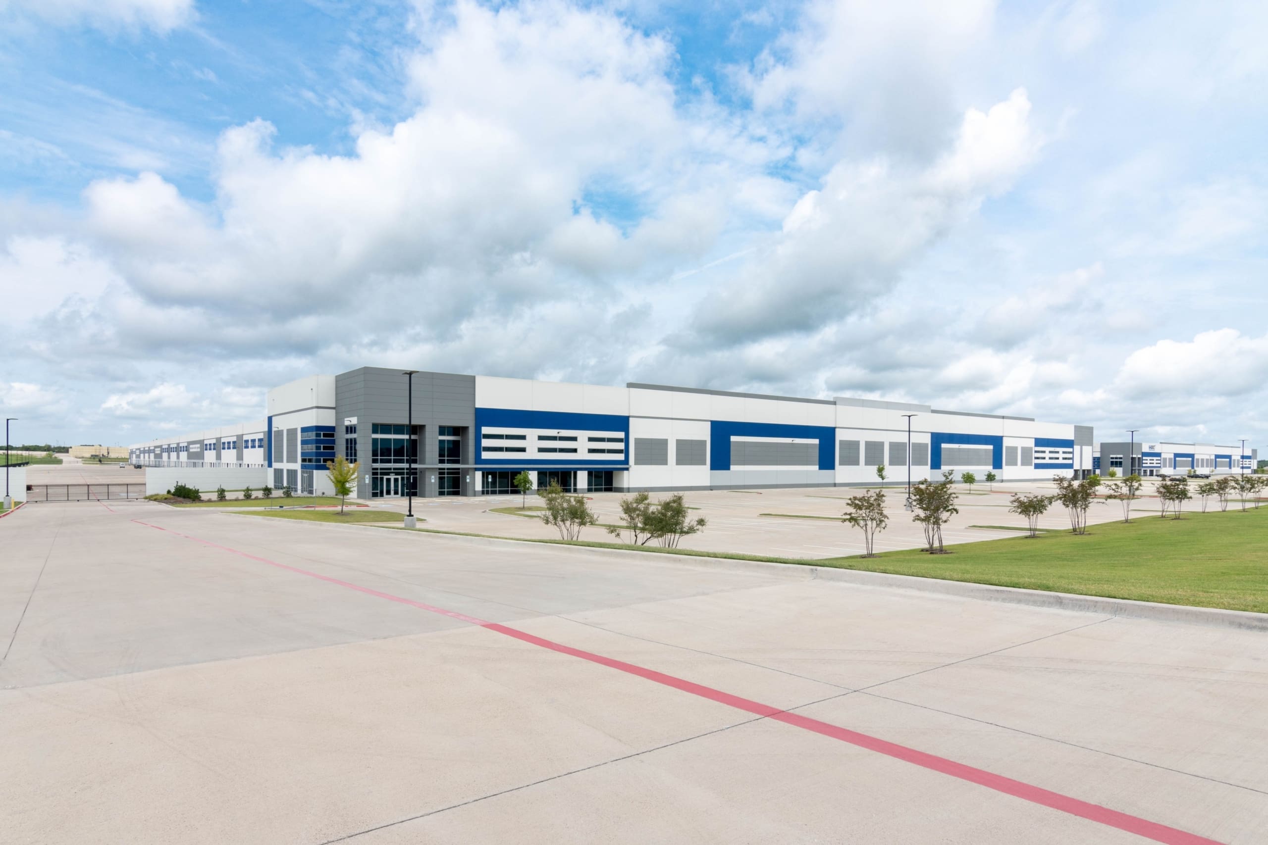 Large modern industrial warehouse with blue and white exterior, empty parking lot, and clear sky with scattered clouds.