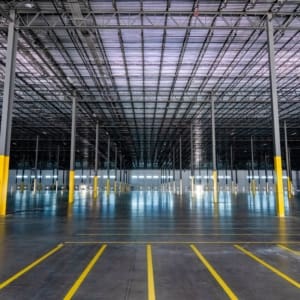 Large, empty warehouse interior with polished concrete floor, yellow safety-marked pillars, and grid-like ceiling structure under bright industrial lighting.