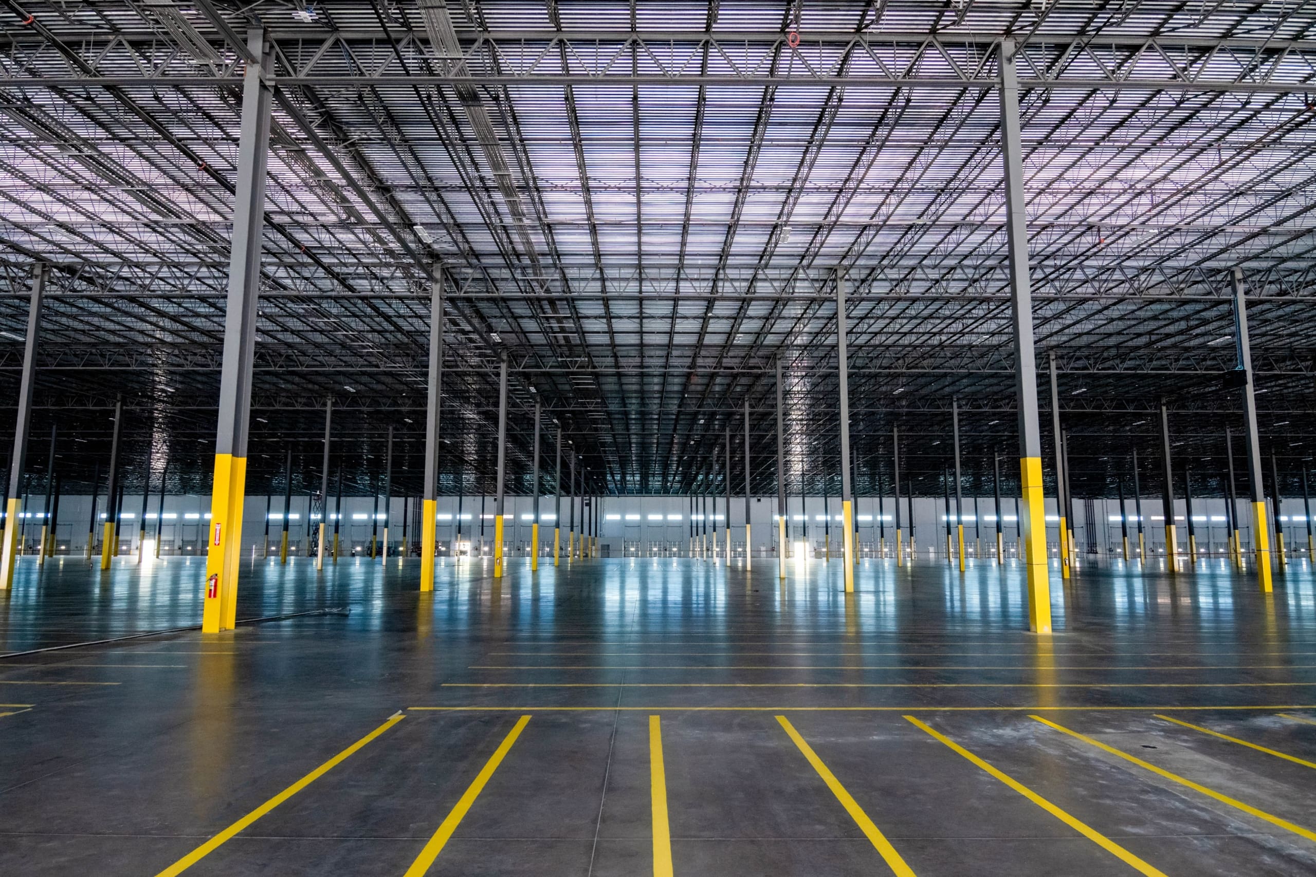 Large, empty warehouse interior with polished concrete floor, yellow safety-marked pillars, and grid-like ceiling structure under bright industrial lighting.
