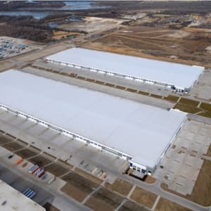 Aerial view of two large, white-roofed warehouse buildings surrounded by parking lots and roads in an industrial area with open fields and water in the background.