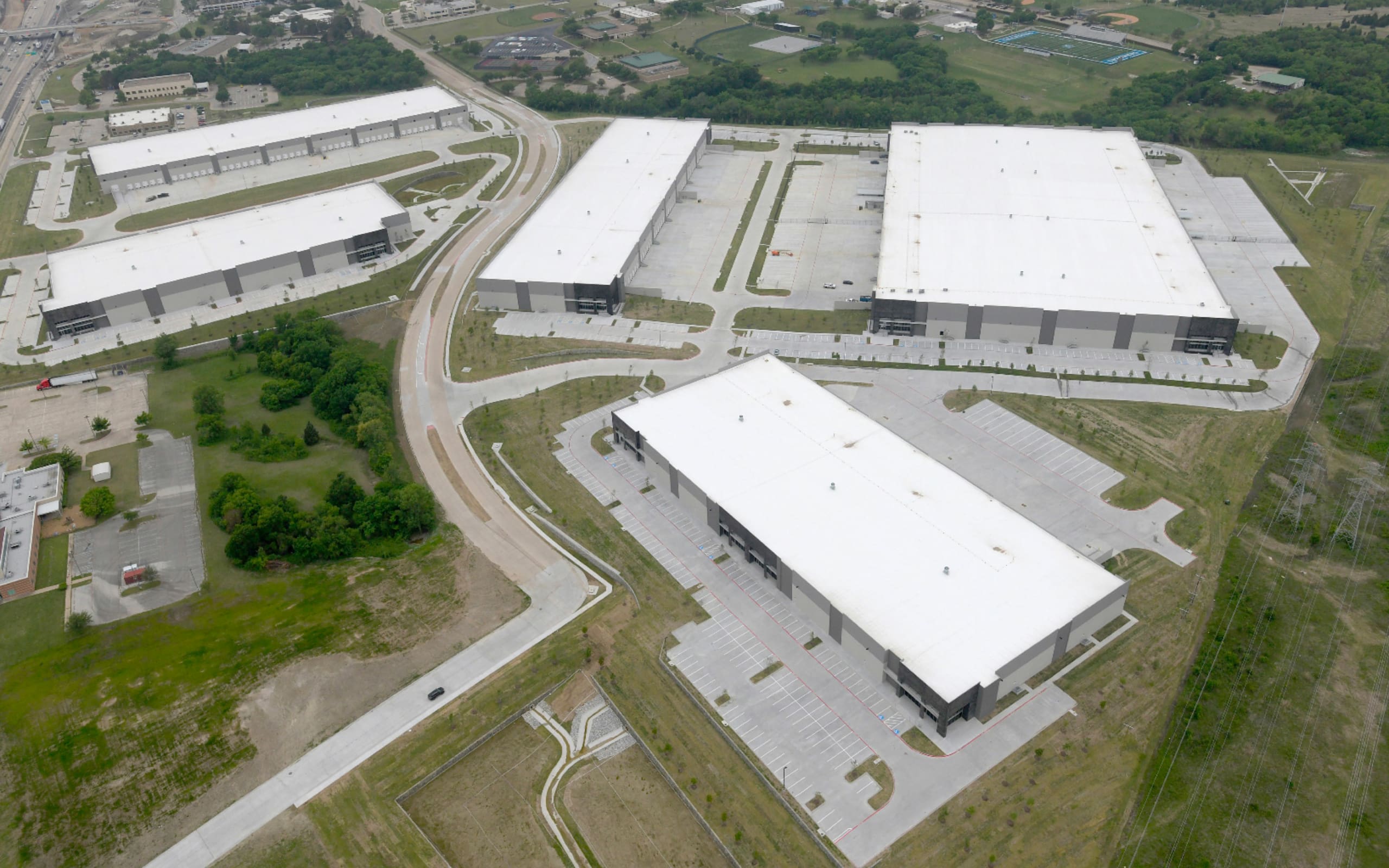 Aerial view of an industrial park with several large warehouse buildings, parking lots, and surrounding green spaces. Roads connect the buildings within the complex.