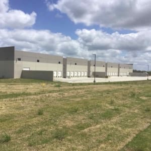 A large, rectangular warehouse building with multiple loading docks is situated in a grassy area under a partly cloudy sky.