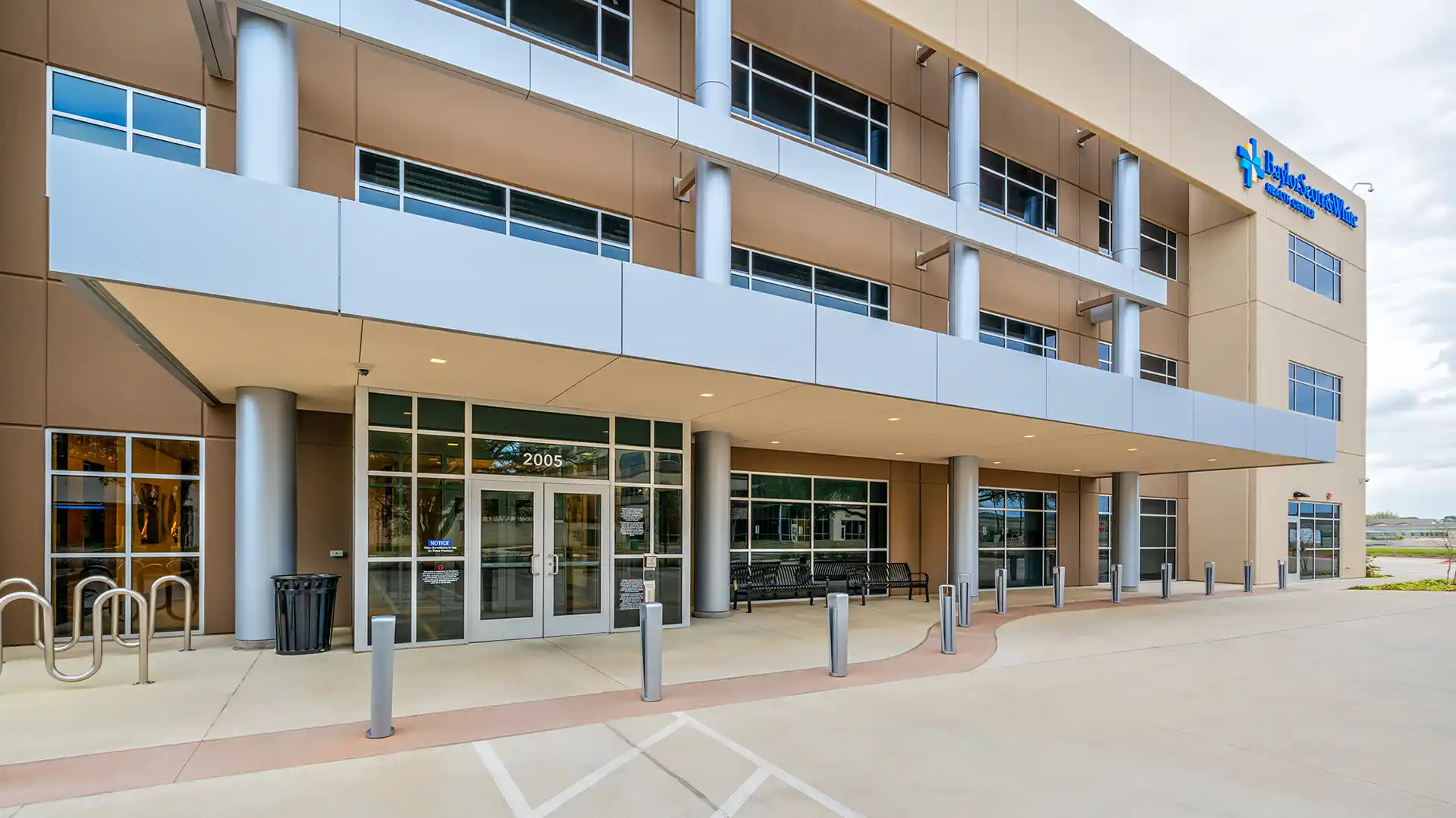 Exterior view of a modern medical building with large glass windows, automatic doors, metal columns, and the Holston Medical Group logo alongside Baylor Scott & White Medical Office Building signage on the upper right corner.