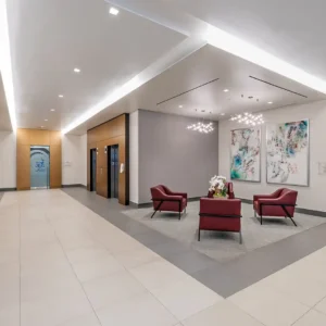 A modern office lobby in the Baylor Scott & White Medical Office Building features three red chairs, two elevators, artwork on the walls, and glass doors labeled "Optometric Specialists.