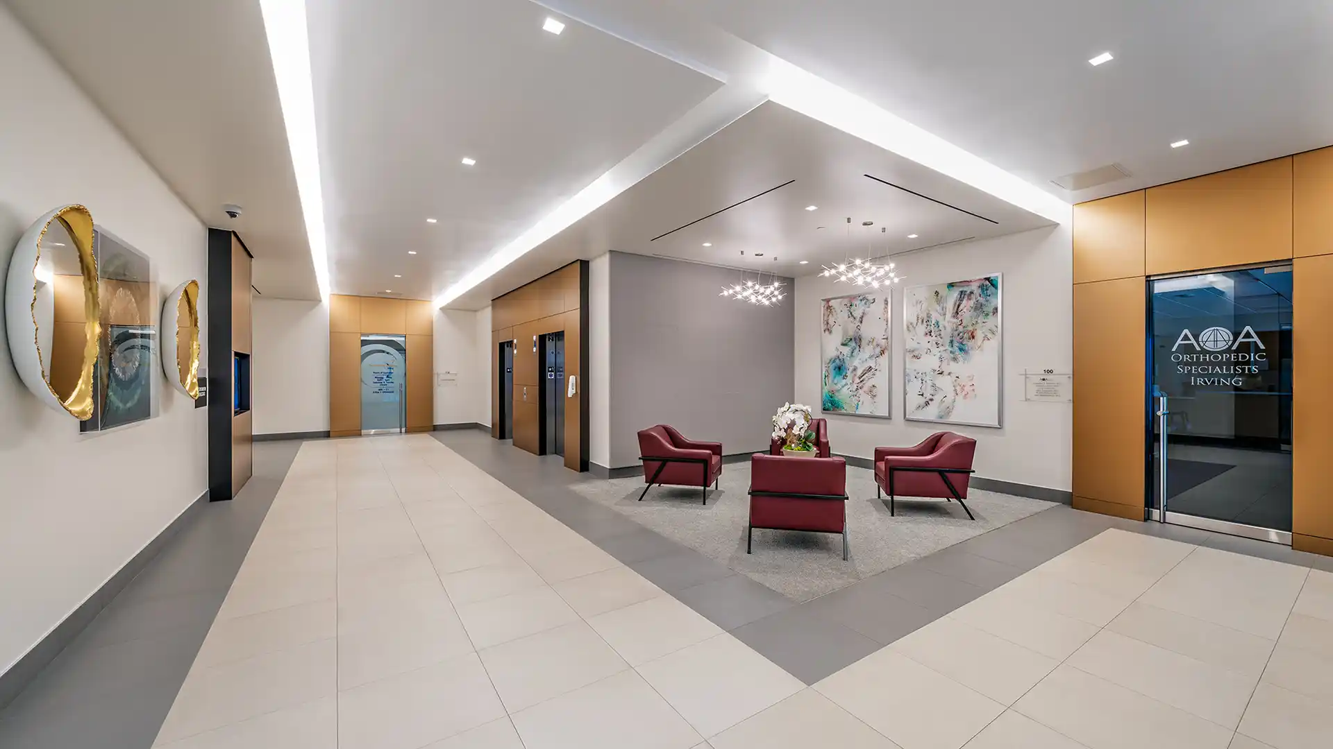 A modern office lobby in the Baylor Scott & White Medical Office Building features three red chairs, two elevators, artwork on the walls, and glass doors labeled "Optometric Specialists.