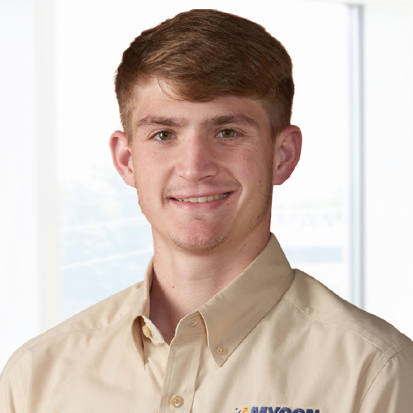 A young man with short brown hair, wearing a beige button-up shirt, smiling in front of a bright background.