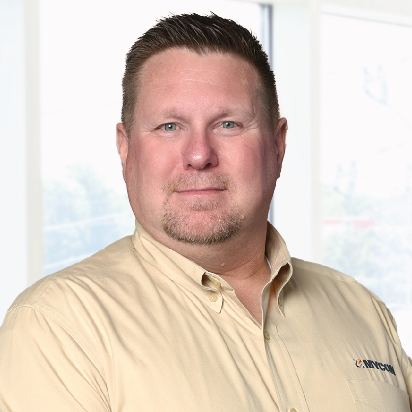 Man wearing a beige button-up shirt standing in front of a window with a soft, natural light background.