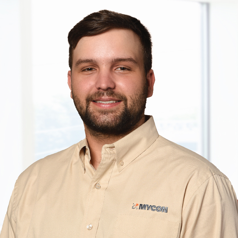 A person with short hair and a beard is smiling. They are wearing a beige shirt with a "MYCON" logo on it, standing in front of a blurred window background.