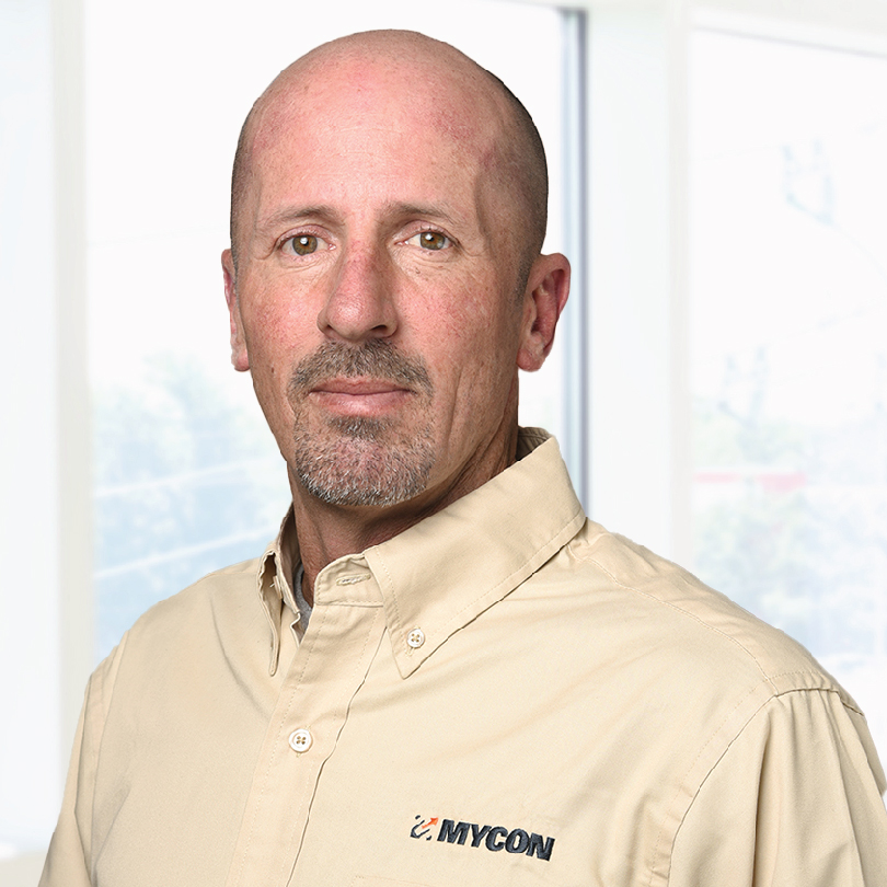 A man with a bald head wearing a beige shirt with "MYCON" logo stands indoors in front of a sunlit window.