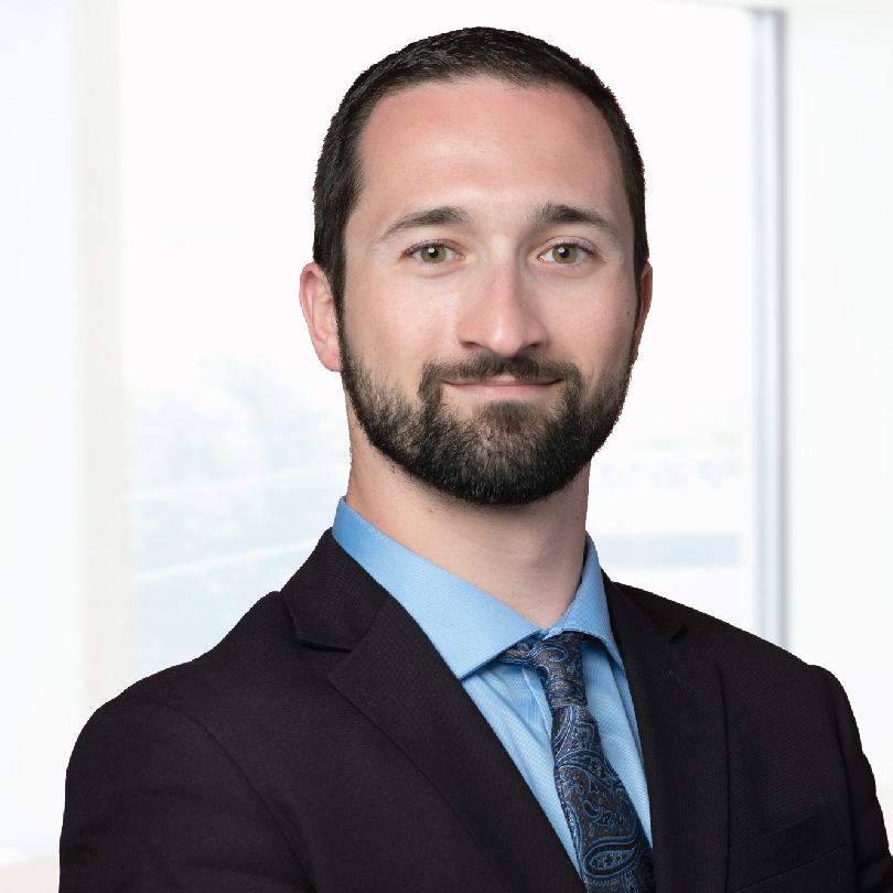 A bearded man in a suit and tie stands in front of a bright office window, looking at the camera with a neutral expression.