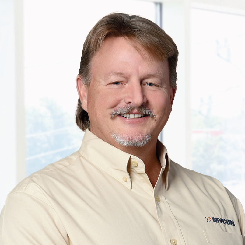 A middle-aged man with light brown hair and a mustache is smiling and wearing a beige button-up shirt in front of a brightly lit window.