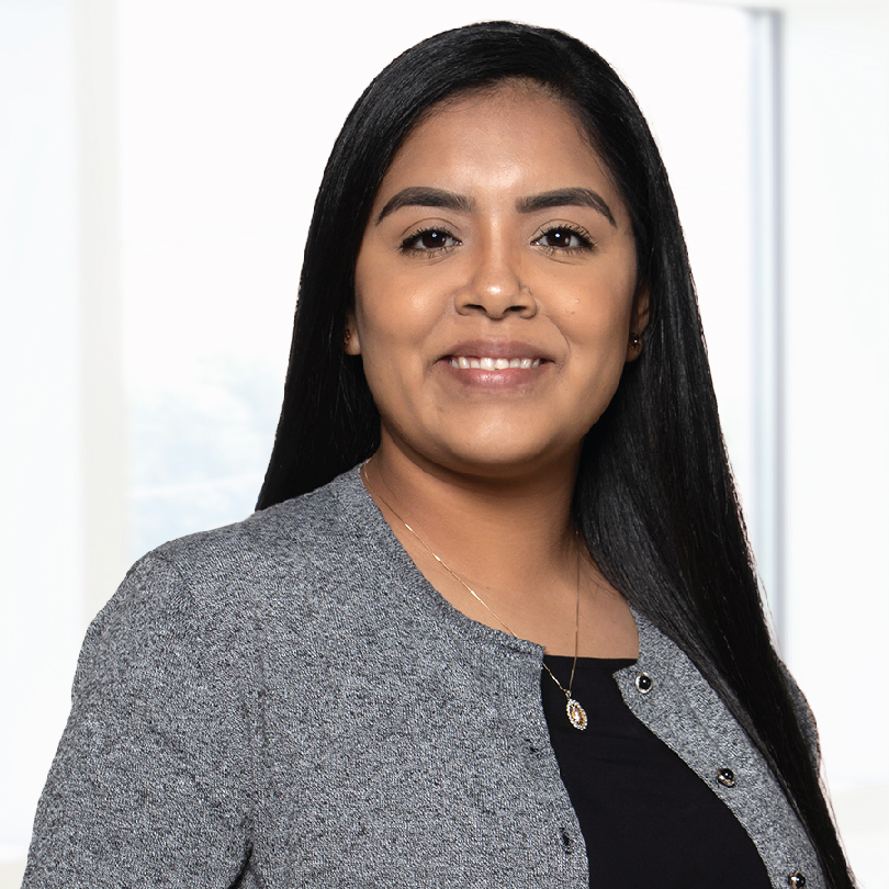A woman with long black hair wearing a grey blazer and black top smiles at the camera against a light background.