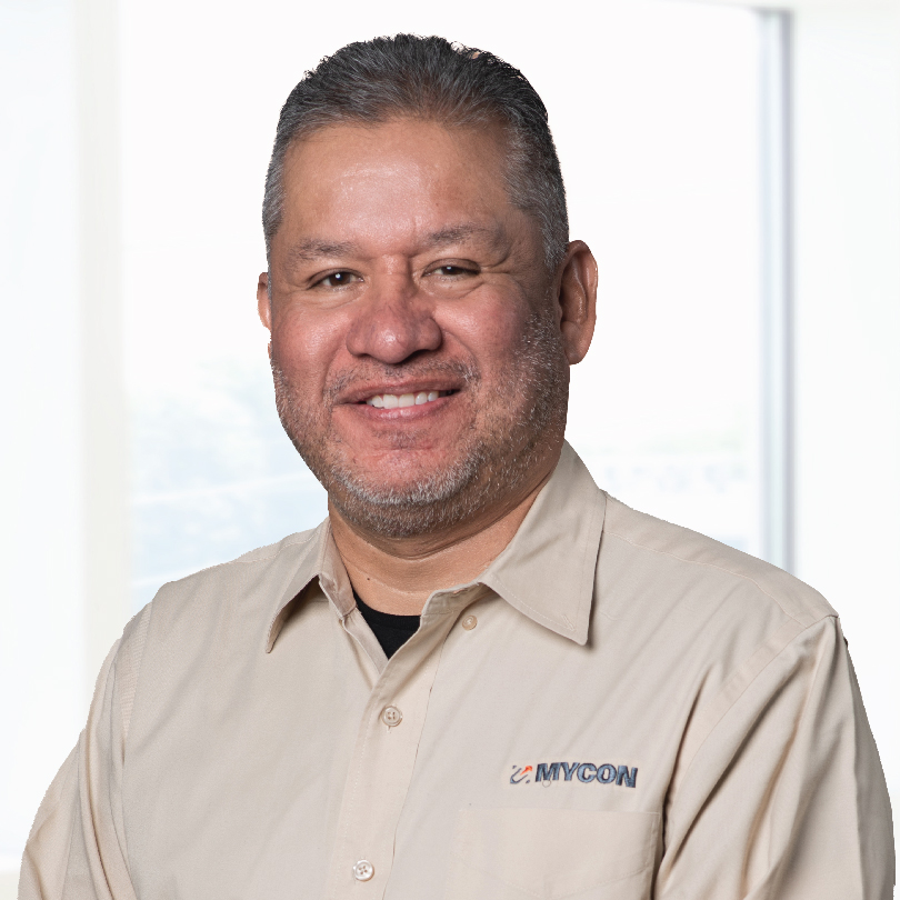 A man with short hair and a beard is smiling. He is wearing a light-colored button-up shirt with a "MYCON" logo on it. The background is an indoor setting with a large window.