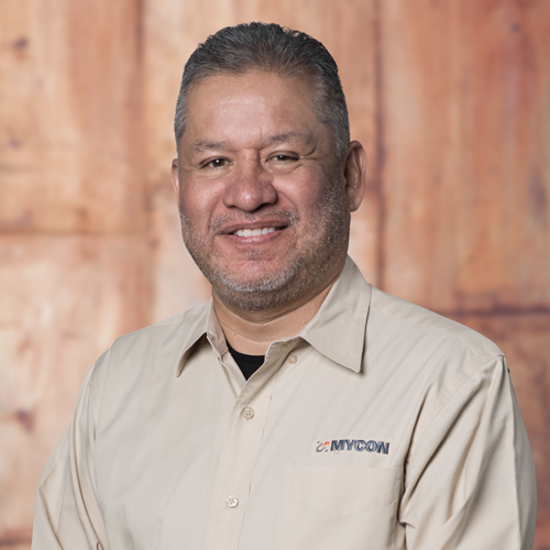 A man in a tan shirt smiling in front of a wooden wall.