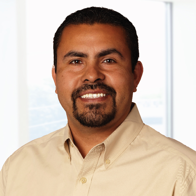A smiling person with a beard and mustache, wearing a beige collared shirt, stands in front of a blurred white and light blue background.
