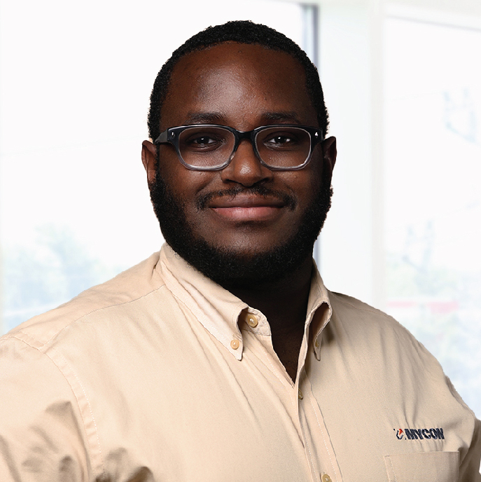 A man wearing glasses and a beige button-down shirt stands in front of a bright window backdrop, looking directly at the camera with a slight smile.