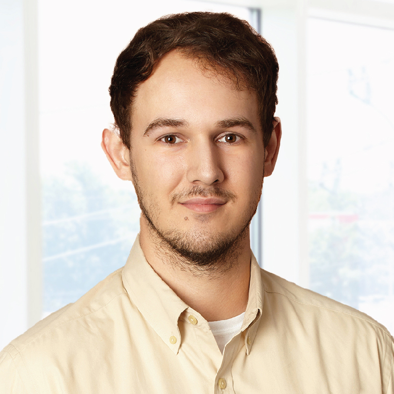 A man with short brown hair and a beard is wearing a beige button-down shirt and standing indoors in front of a window.