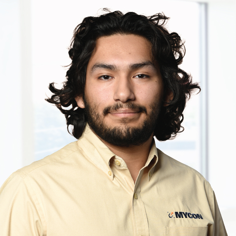 A person with long, curly hair and a beard is wearing a beige shirt with a logo that reads "MYCOM". They are posing in front of a blurred window background.