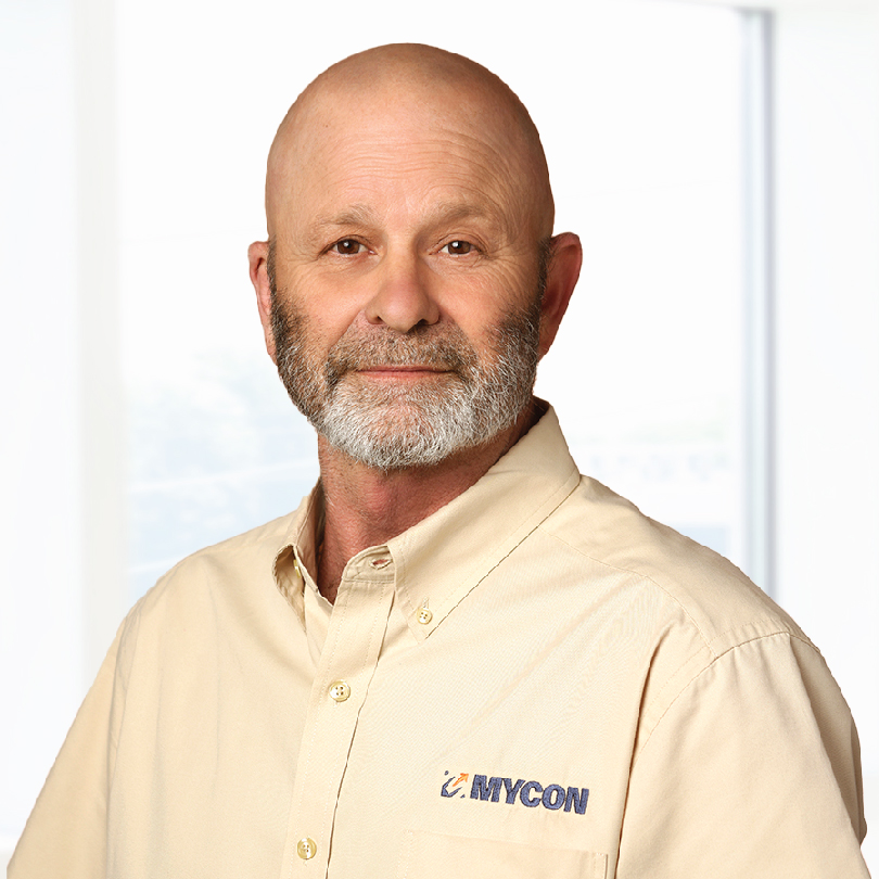 A man with a bald head and gray beard wearing a beige shirt with a "MYCON" logo stands in front of a bright background.