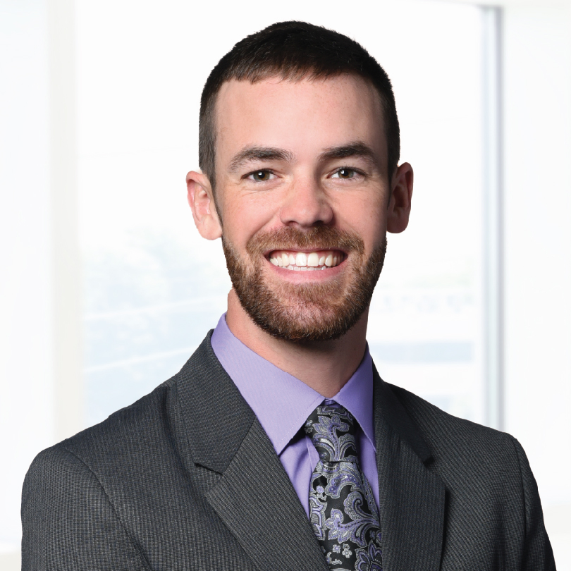 A man with short dark hair, a beard, and a wide smile is wearing a grey suit with a lavender shirt and patterned tie. He is standing against a blurred light background.