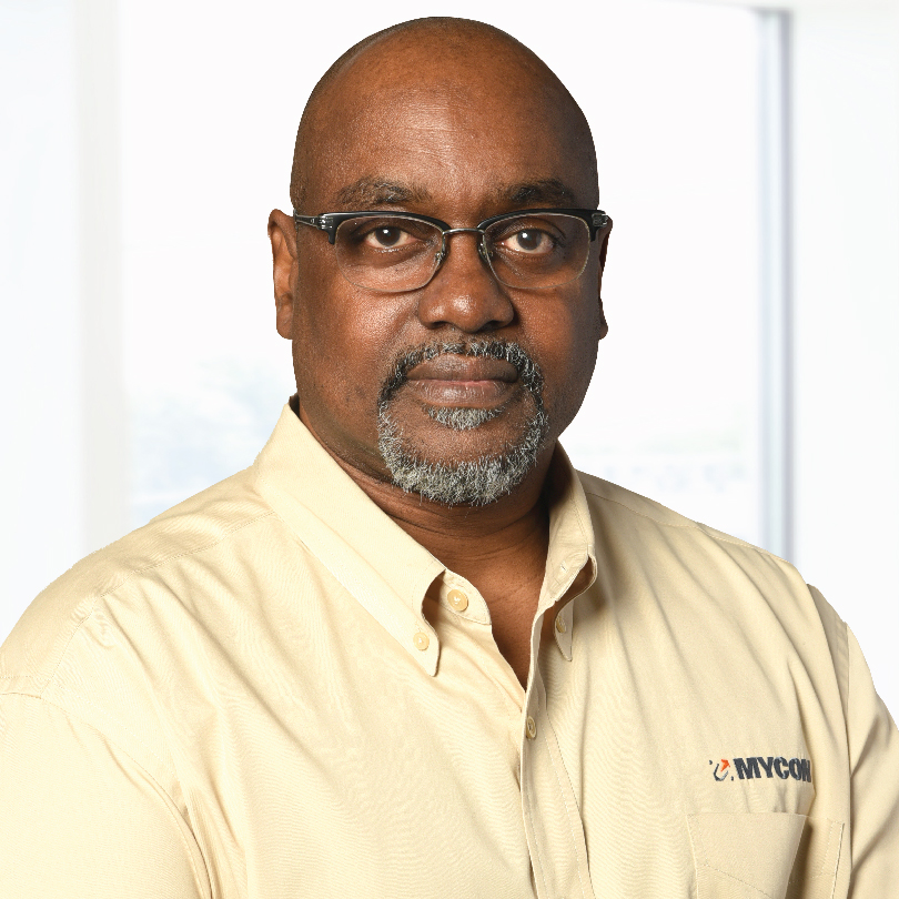 A man with glasses and a goatee, wearing a beige collared shirt with a "MYCOM" logo, stands in front of a light background.