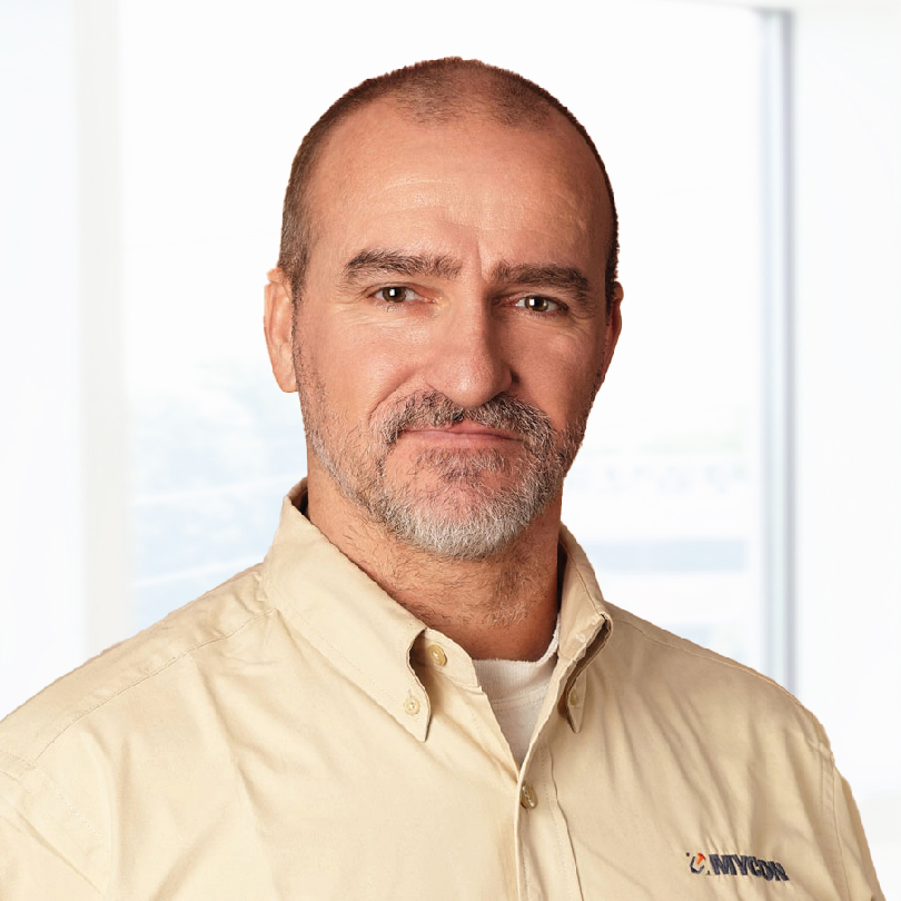 A man with a shaved head and short beard looks directly at the camera. He is wearing a beige button-up shirt with a company logo on the chest. The background shows a bright, blurred interior.