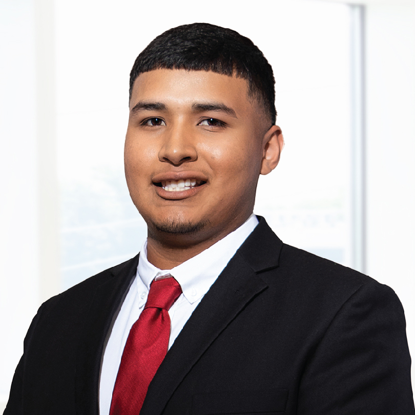 A young man in a black suit with a white shirt and red tie smiles while posing for a professional photo, with a light-colored background.