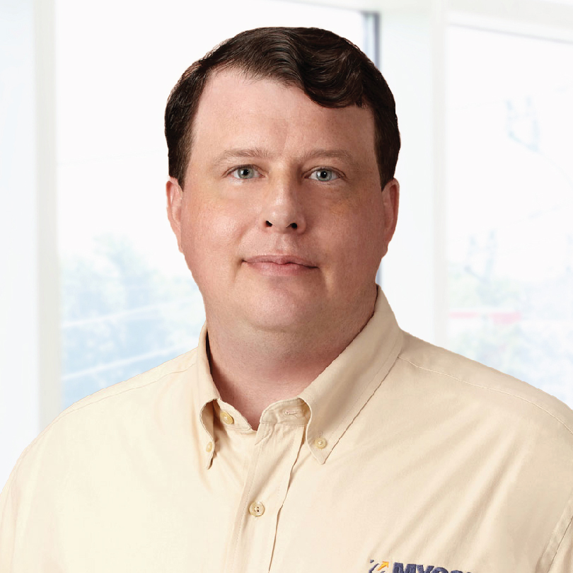 Middle-aged man with short brown hair and a neutral expression, wearing a beige collared shirt with a partial logo visible. Background shows an office setting with large windows.