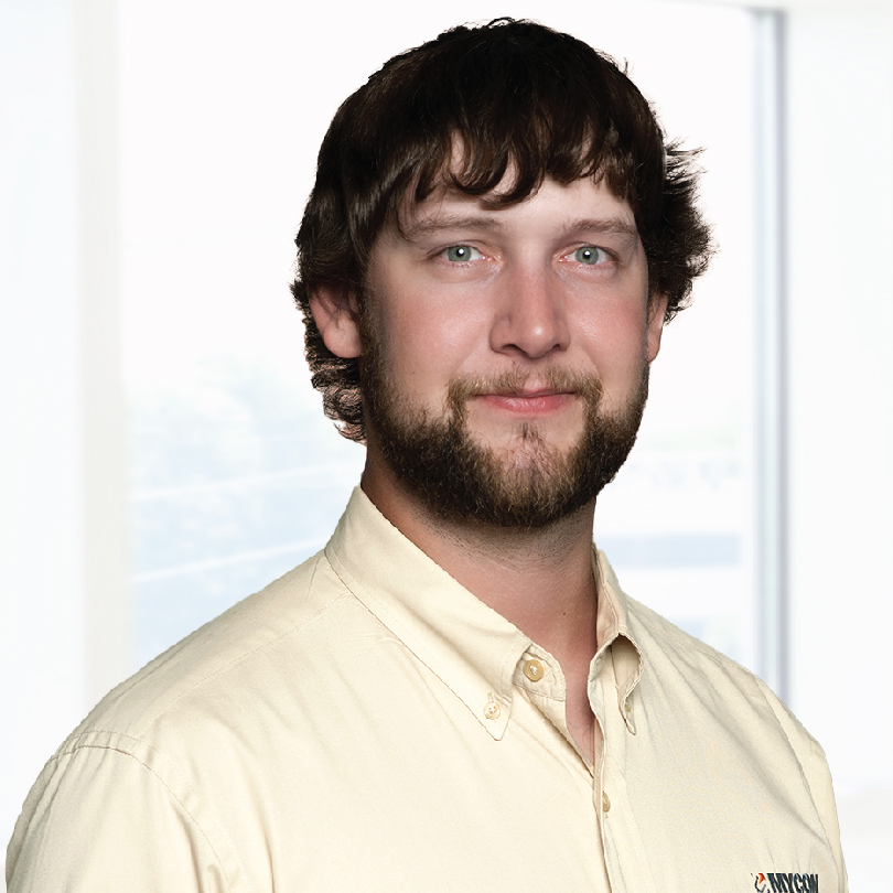 A person with short brown hair and a beard is wearing a beige button-up shirt. They are standing in front of a bright window.