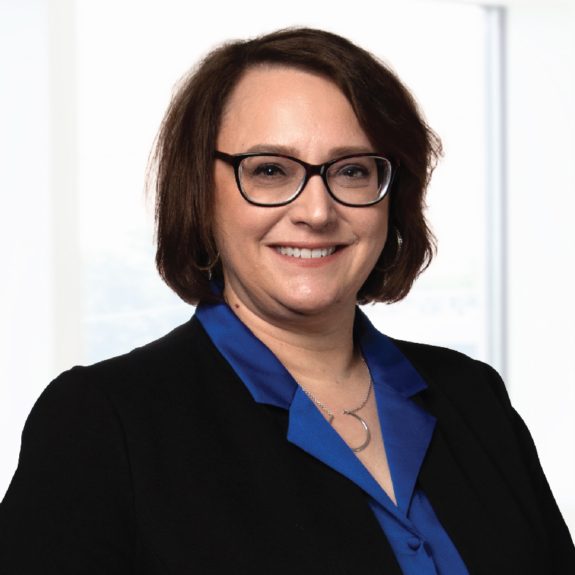 A woman with short brown hair and glasses is wearing a blue blouse and black blazer, standing in front of a white background.